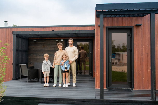 Heureuse famille contemporaine de jeunes parents et deux petits enfants debout sur le patio de leur nouvelle maison sur fond de fauteuils noirs - Photo, image
