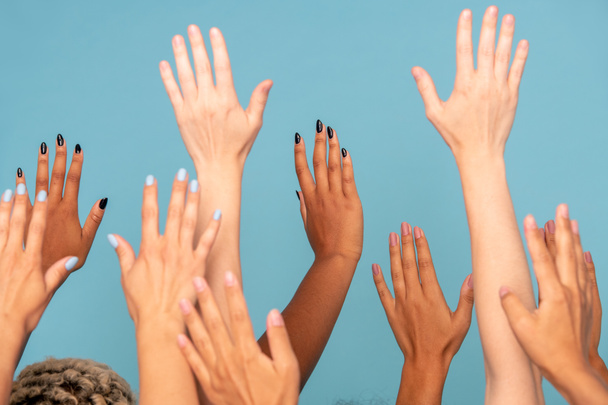 Mains d'un grand groupe de jeunes femmes de différentes ethnies à la peau blanche et foncée levant les bras devant la caméra sur fond bleu - Photo, image
