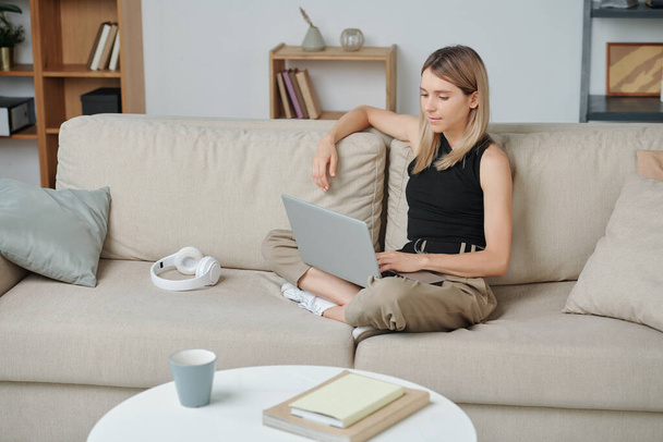 Χαλαρός έφηβος σε casualwear κάθεται σε μαλακό άνετο καναπέ με φορητό υπολογιστή μπροστά και βλέποντας ταινία ή surfing στο διαδίκτυο κατά την αναψυχή - Φωτογραφία, εικόνα