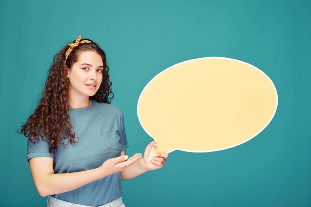 Smiling student girl with curly hair holding speech bubble banner while making presentation against blue background - Photo, Image