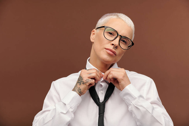 Young female with short dyed blond hair, piercing in nose and tattoos on right hand doing buttons of white shirt and looking at you - Photo, Image