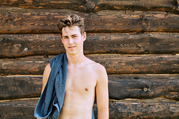 Young topless sportsman with blue towel on shoulder standing against wooden wall in front of camera after outdoor sports training - Fotografie, Obrázek