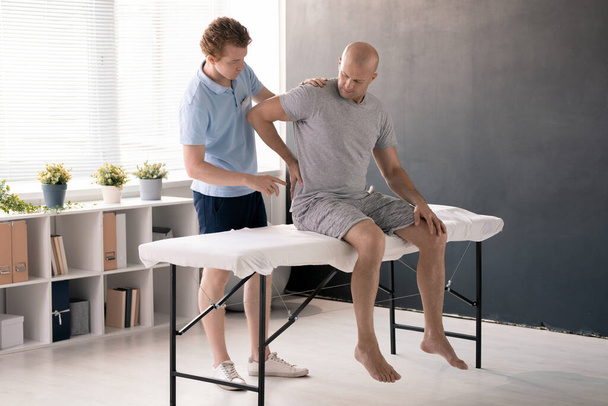 Dojrzały mężczyzna dotyka bolesnych pleców podczas siedzenia na kanapie medycznej w klinikach rehabilitacyjnych i konsultacji z fizjoterapeutą - Zdjęcie, obraz