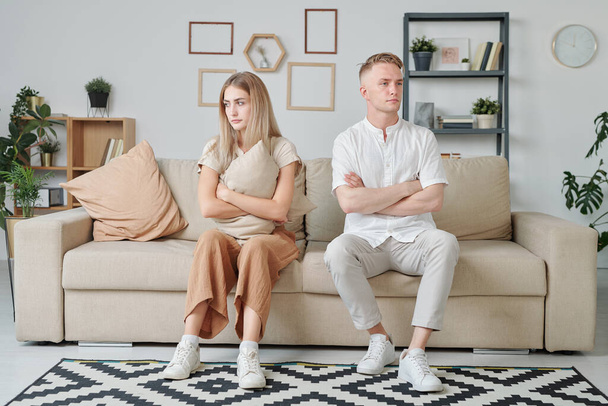 Молодая супружеская пара со скрещенными руками и оскорбленным выражением лица, сидящая на диване после ссоры или ссоры и хранящая молчание - Фото, изображение