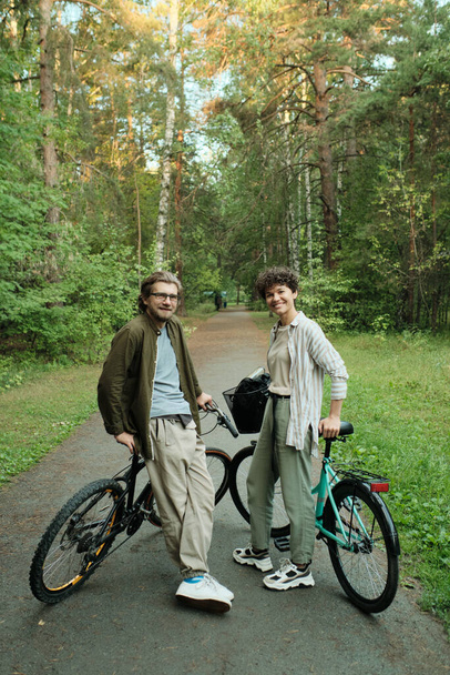 Young joyful couple with bicycles standing on road between green trees in front of camera and looking at you while enjoying warm day - Photo, Image