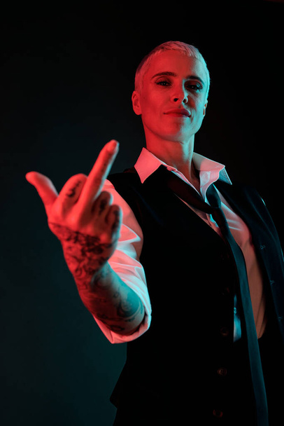 Joven rubia en ropa formal mostrando gesto grosero mientras está de pie en la oscuridad y manteniendo el brazo con tatuajes delante de la cámara - Foto, imagen