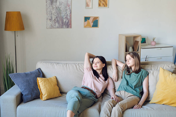Junge Mutter und Tochter relaxen auf Couch im Wohnzimmer gegen Wand mit Strumpfhosen, Lampe, grüner Zimmerpflanze und ein paar Möbeln - Foto, Bild