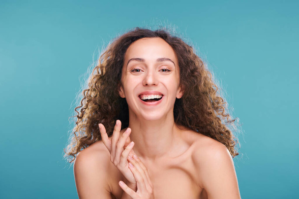Fröhliche junge brünette Frau mit gesunden Zähnen lacht vor der Kamera, während sie ihre Hände vor blauem Hintergrund dicht vor dem Gesicht hält - Foto, Bild