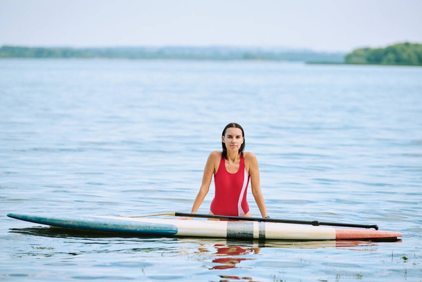gelukkig jong brunette vrouw in rood badpak staande in water door surfplank terwijl genieten van eenzaamheid en zomervakantie in resort - Foto, afbeelding