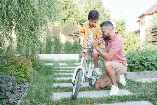 Ragazzino seduto in bicicletta in attesa che suo padre fissi l'altezza del manubrio all'aperto nella soleggiata giornata estiva - Foto, immagini