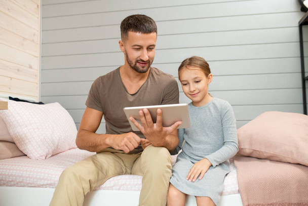 Mladý usmívající se otec s digitálním tabletem sedí na posteli vedle své roztomilé dcerušky, zatímco oba sledují film nebo procházejí zprávy - Fotografie, Obrázek