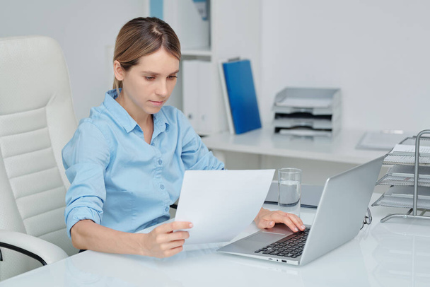 Занят молодой элегантный секретарь или бухгалтер, читающий финансовую бумагу и вводящий данные в компьютерную базу, сидя в кресле за столом - Фото, изображение