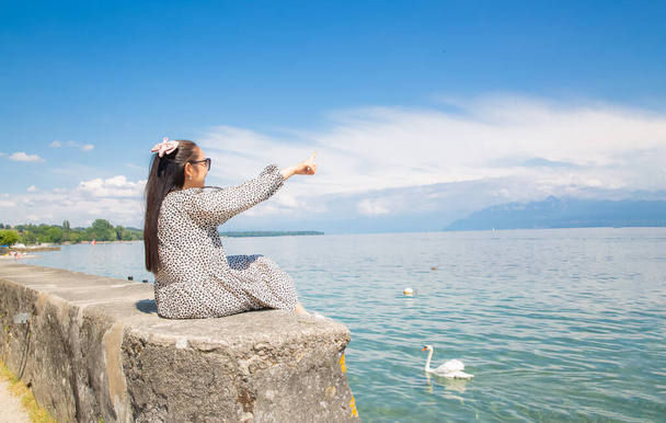 Όμορφη Ασιάτισσα με λευκό δέρμα και μακριά μαύρα μαλλιά με μαύρα γυαλιά. να κάθεσαι πάνω σε πέτρα και να βλέπεις τη θέα στη λίμνη σε μια καθαρή μέρα στον ουρανό, τον τουρισμό και τις καλοκαιρινές διακοπές. - Φωτογραφία, εικόνα