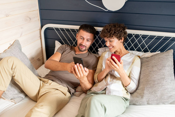 Молодий чоловік показує свою щасливу дружину цікаве онлайн-відео або пост в соціальних мережах в смартфоні, одночасно відпочиваючи на ліжку на дозвіллі
 - Фото, зображення