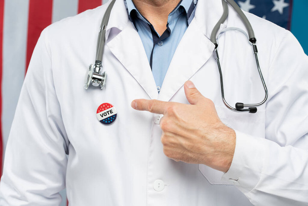 Hand van hedendaagse Amerikaanse burger in doktersuniform wijzend naar insignes op zijn borst, wat betekent dat hij gaat stemmen. - Foto, afbeelding