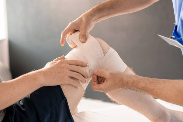 Mains du clinicien enveloppant le genou droit du patient avec un bandage flexible pendant la procédure médicale avant l'entraînement physique de réadaptation - Photo, image