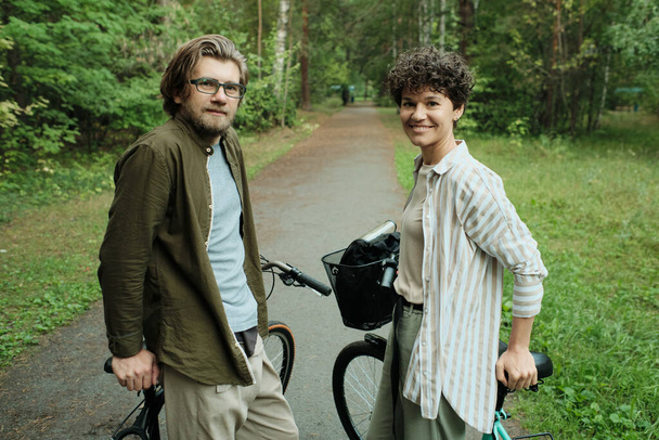 Молодая пара велосипедистов в повседневной одежде стоит на лесной дороге между зелеными деревьями и смотрит на вас, наслаждаясь летними выходными - Фото, изображение