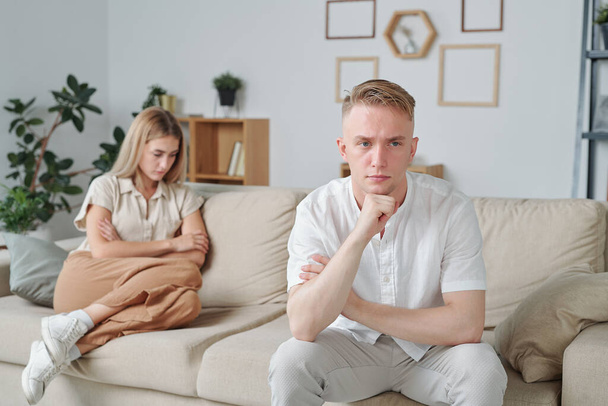 Young tense man with upset facial expression sitting on couch on background of his unhappy wife with her arms crossed by chest - Fotoğraf, Görsel