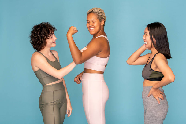Happy young African woman showing strength while Caucasian girl in tracksuit touching her muscles and Asian female laughing at them - Photo, Image