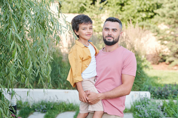Horizontal medium portrait of bearded man standing outdoors holding his son looking at camera - Photo, Image