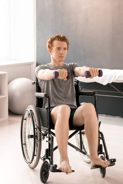 Mladý sportovec v aktivním oblečení sedí na invalidním vozíku a natahuje paže s činkami dopředu během rehabilitačního kurzu na klinikách - Fotografie, Obrázek