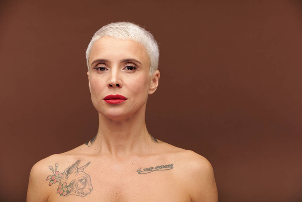 Mature woman with short blond hair, tattoos on neck and chest and red lipstick on lips looking at you while standing in front of camera - Photo, Image