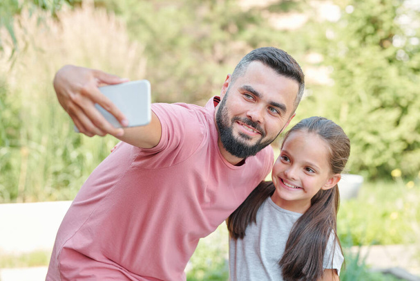 Moderno jovem pai adulto vestindo camiseta rosa pálida tomando selfie tiro com sua filha, tiro médio horizontal - Foto, Imagem