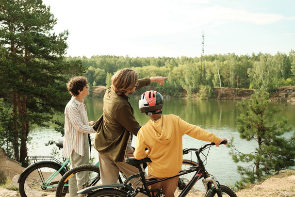 Joven en ropa casual apuntando a los árboles en la orilla opuesta del río mientras muestra a su hijo adolescente donde es realmente genial montar en bicicleta - Foto, Imagen