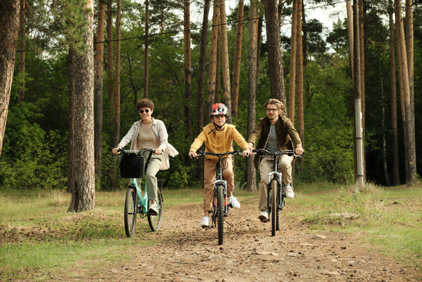 Happy active family of young husband, wife and son riding bicycles along forest path against pinetree trunks and other trees on summer day - Photo, Image