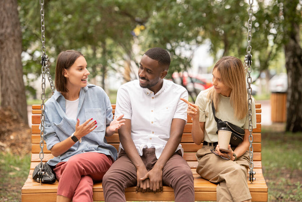 Happy young African man sitting on wooden swing between two girls having argument and looking at one of them during conversation - Valokuva, kuva