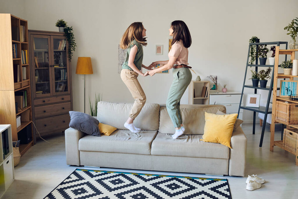 Joyeux jeune femme et sa fille adolescente aux cheveux blonds sautant ensemble sur un grand canapé dans le salon tout en tenant par les mains - Photo, image