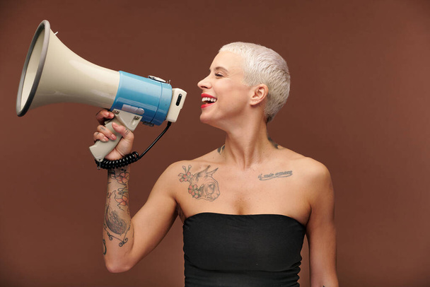 Щаслива молода блондинка з татуюваннями на руці, грудях і шиї і мегафон в руці робить важливе оголошення в ізоляції
 - Фото, зображення