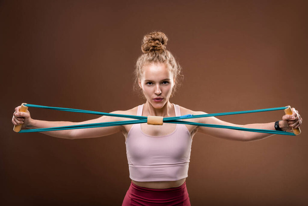 Young active female in tracksuit stretching elastic band in front of herself and looking at you while exercising in front of camera in isolation - Foto, Imagem
