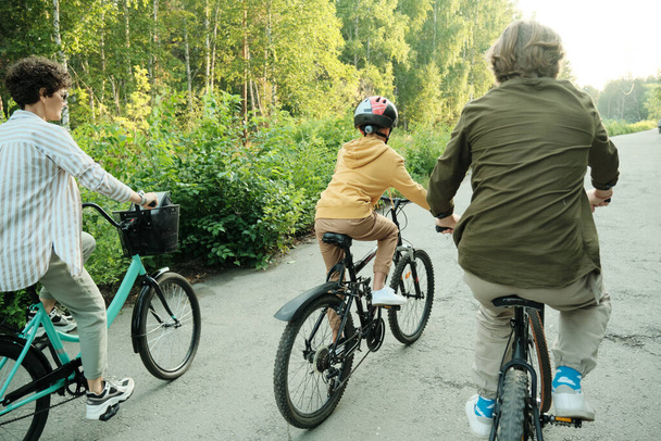 Ενεργός οικογένεια του συζύγου, της συζύγου και του γιου τους της στοιχειώδους ηλικίας ιππασία ποδήλατα κατά μήκος του δρόμου στο σύγχρονο πάρκο την ηλιόλουστη μέρα ή το πρωί - Φωτογραφία, εικόνα