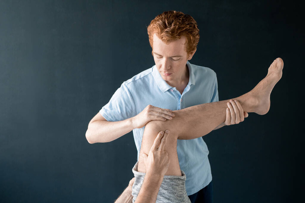 Профессиональный клиницист мужской реабилитации, держащий ногу пациента на медицинском диване во время физических упражнений и помогающий ему согнуть ее - Фото, изображение