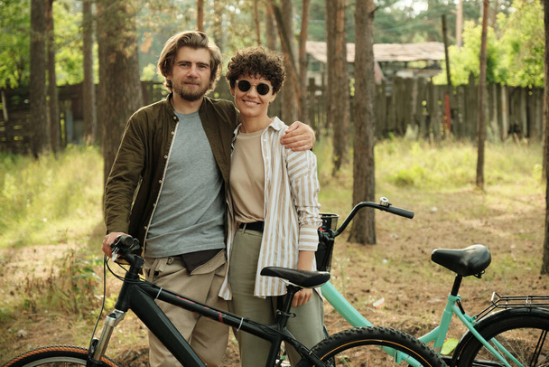 Νεαρός στοργικός άντρας αγκαλιάζει την ευτυχισμένη του γυναίκα με γυαλιά ηλίου ενώ και οι δύο στέκονται μπροστά στην κάμερα δίπλα στα ποδήλατά τους στο αγροτικό περιβάλλον - Φωτογραφία, εικόνα