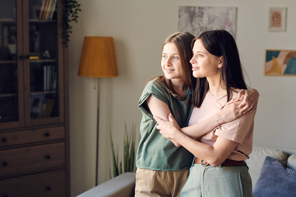 Two young pretty females with blond and dark long hair standing in embrace against wall with paintings and lamp in living-room - Fotografie, Obrázek