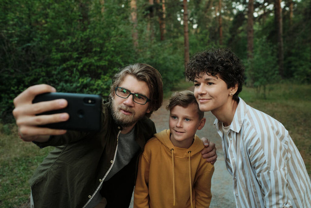 Família contemporânea de pais felizes e seu filho de pé perto uns dos outros no caminho da floresta ou estrada e fazer selfie - Foto, Imagem