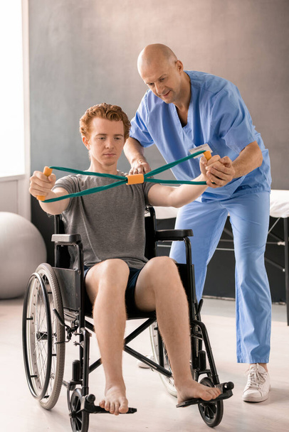 Médico maduro em uniforme azul ajudando o jovem paciente em cadeira de rodas enquanto apoia a mão durante o exercício físico com banda de resistência - Foto, Imagem