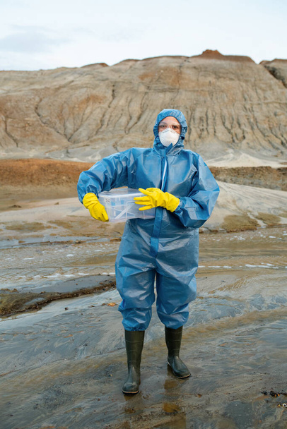 Jonge vrouwelijke wetenschapper in beschermende overall, masker, rubber handschoenen en laarzen met plastic container met water en bodem monsters - Foto, afbeelding