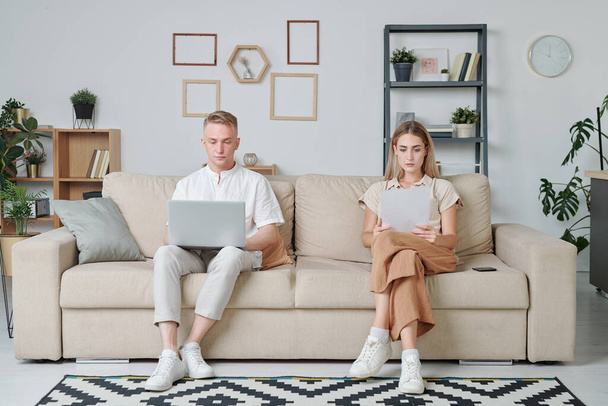 Twee jonge studenten zitten op de bank in de woonkamer, terwijl meisje kijkt door de informatie in de kranten en man die online gegevens leest - Foto, afbeelding