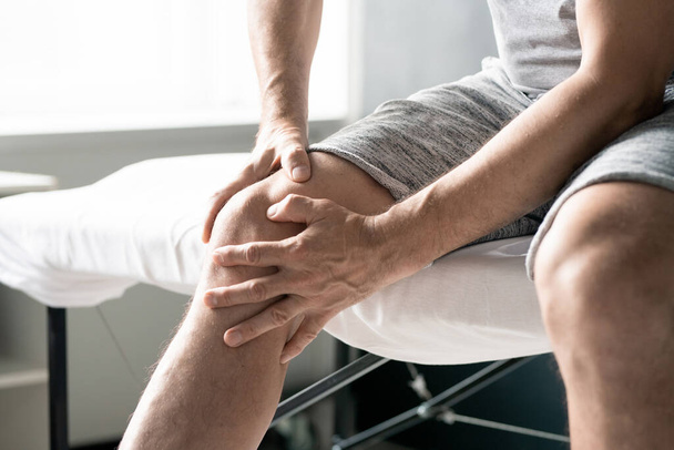 Handen van de hedendaagse volwassen man masseren zijn rechterknie tijdens het zitten op tafel voor fysiotherapeutische procedures in klinieken of medisch kantoor - Foto, afbeelding
