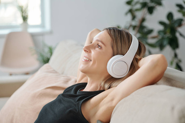 Side view of young blond smiling woman in headphones enjoying music while lying on back of couch in home environment and having rest - Zdjęcie, obraz