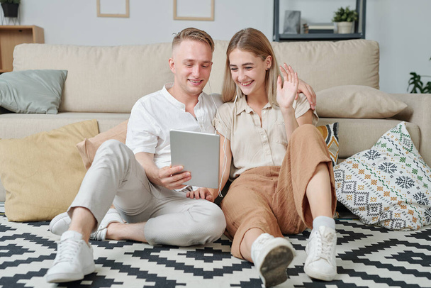 Cheerful young restful couple in earphones sitting on the floor by couch and talking to friends or parents through video chat in living-room - Foto, imagen