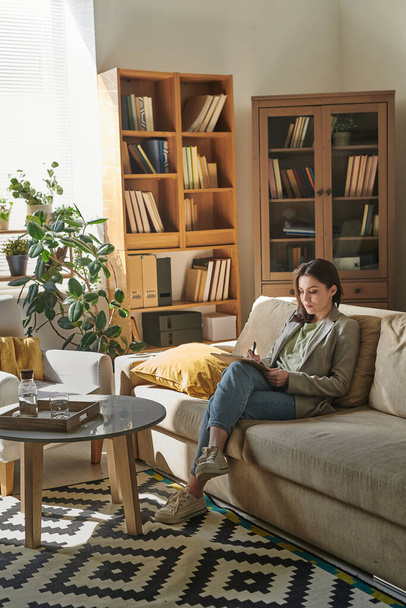 Fiatal nő ül kényelmes kanapén a modern irodai szobában kezében írótábla jegyzetel - Fotó, kép
