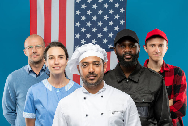 Gruppo multiculturale di giovani uomini e donne contemporanei di varie professioni in uniforme contro la bandiera americana - Foto, immagini