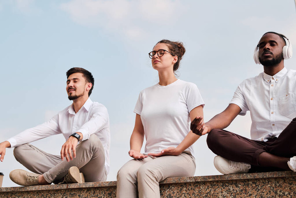 Gruppe junger multiethnischer Menschen sitzt in entspannter Position und meditiert, um sich auf Gedanken im Freien zu konzentrieren - Foto, Bild