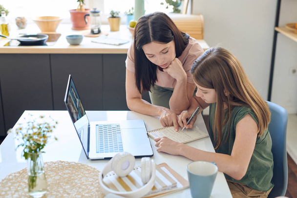 Young brunette female in casualwear pointing at notes of teenage daughter in notepad while helping her with home assignment - 写真・画像