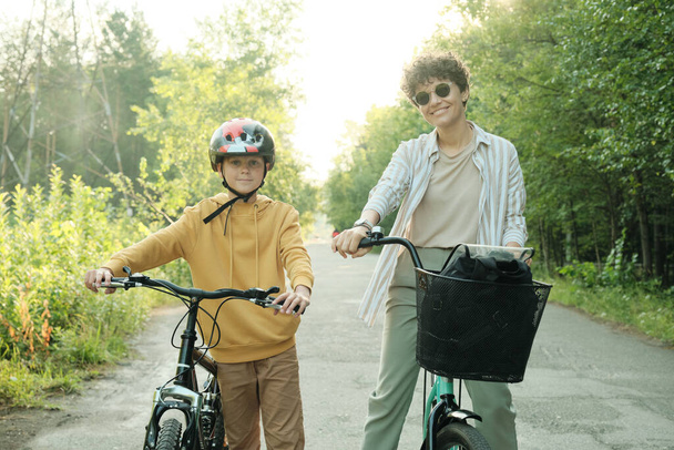 Νεαρή χαρούμενη γυναίκα και ο γιος της σε στοιχειώδη ηλικία με προστατευτικό κράνος και κίτρινη κουκούλα να κάνουν περίπατο στα ποδήλατα την καλοκαιρινή μέρα - Φωτογραφία, εικόνα