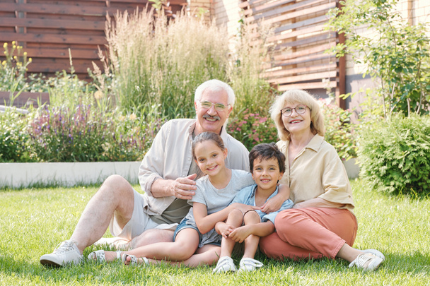 Груповий портрет веселої бабусі, дідуся і двох онуків, які сидять разом на газоні на подвір'ї, дивлячись на камеру посміхаючись
 - Фото, зображення